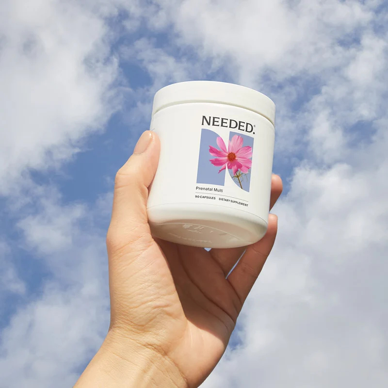 A hand holds a white container labeled "NEEDED:" with a pink flower on the label, against a partly cloudy sky background.