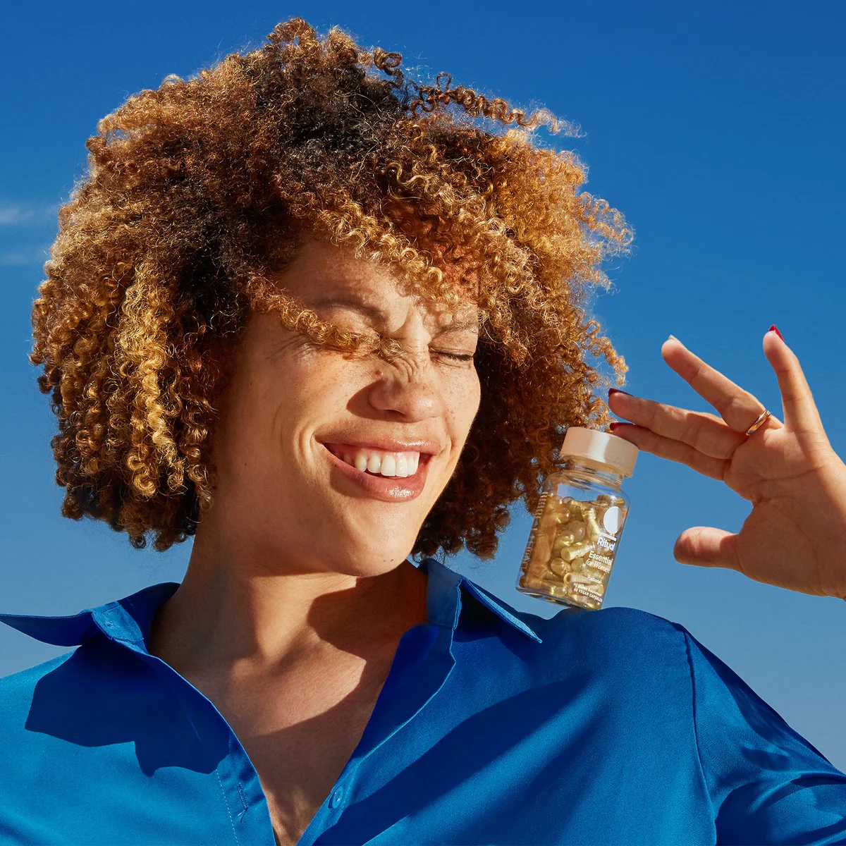 Smiling person in a blue shirt holds a small bottle of capsules against a bright blue sky, squinting one eye in the sunlight.