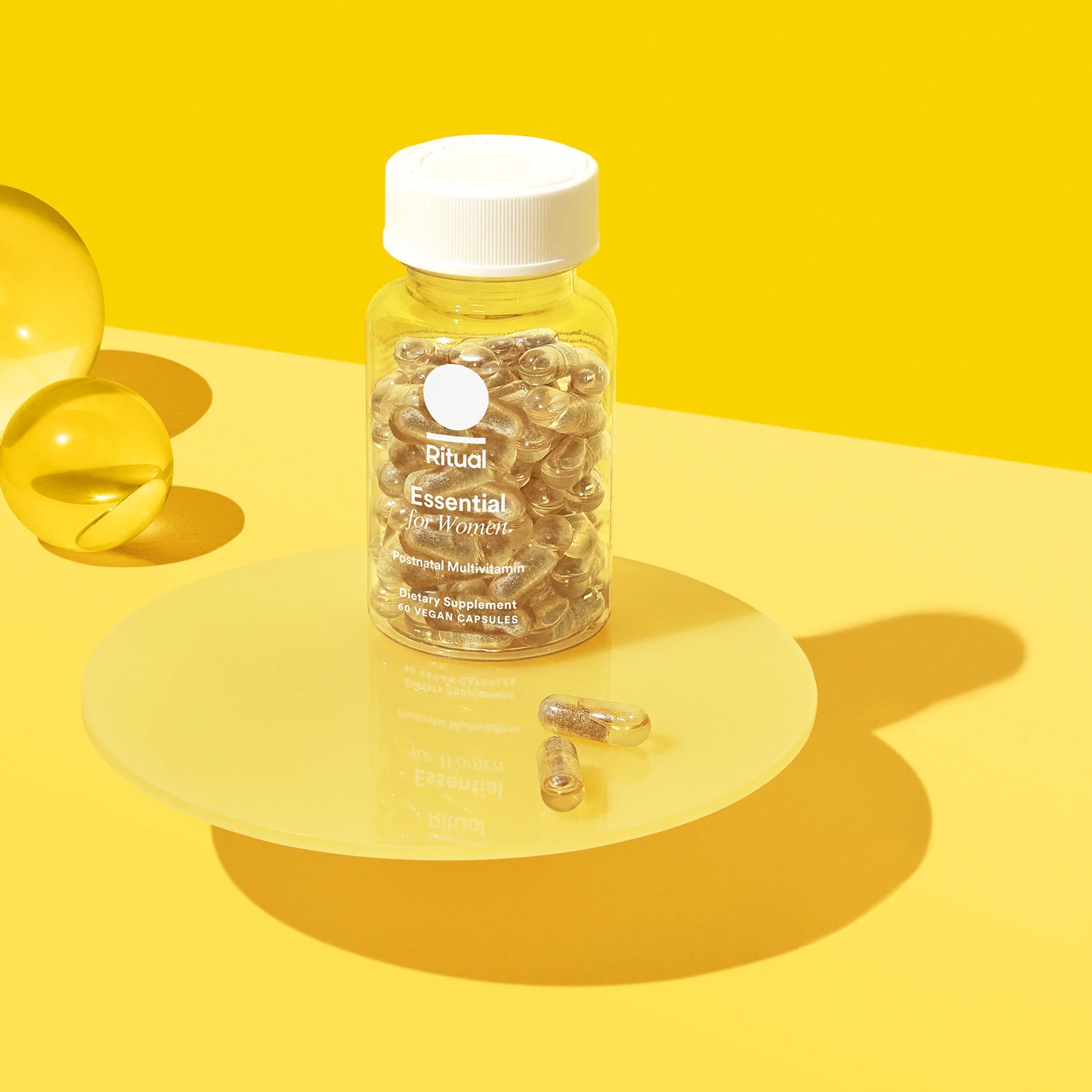 A clear bottle labeled "Ritual Essential for Women" multivitamins sits on a yellow surface with two capsules beside it and a glass sphere in the background.