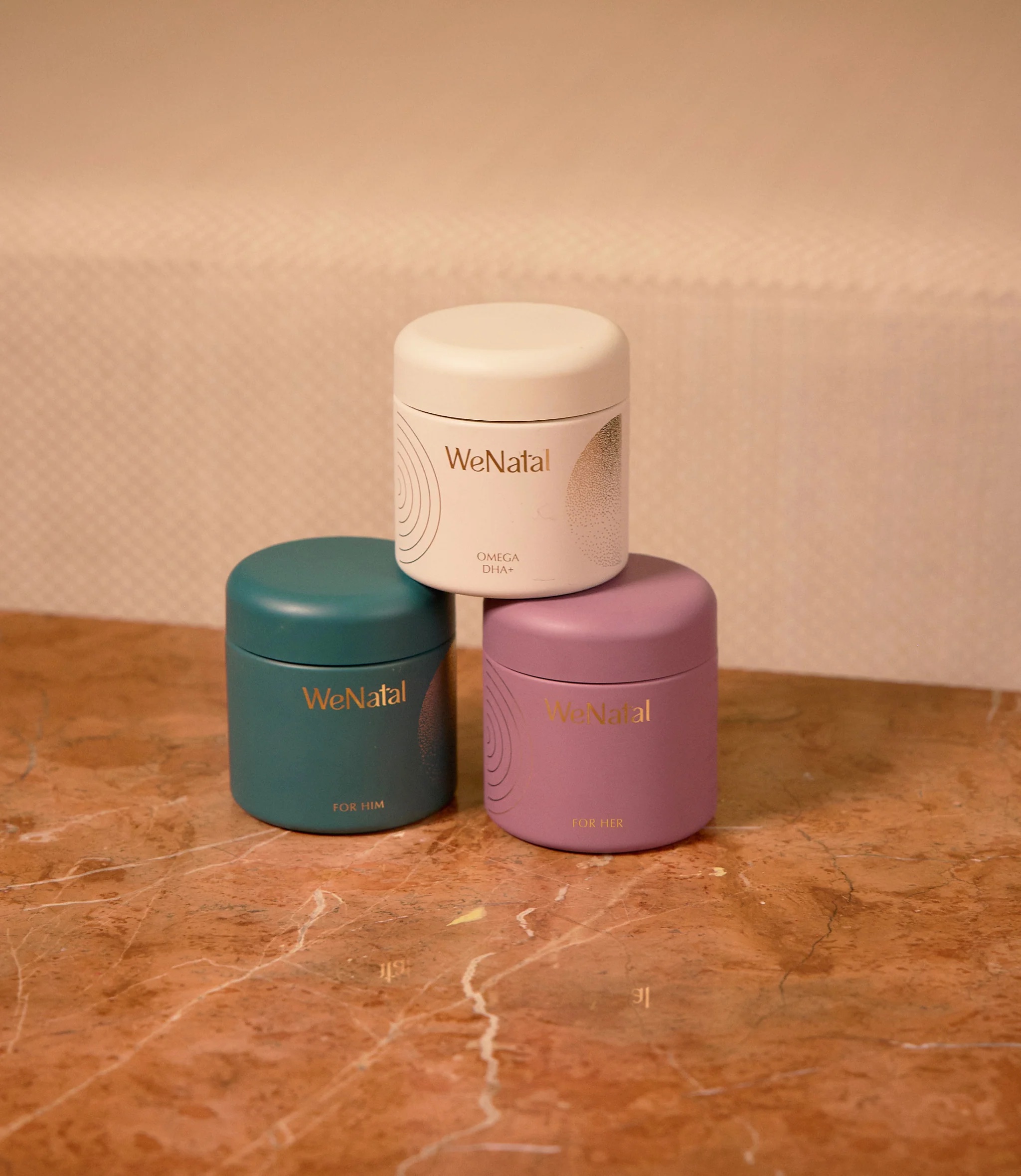 Three WeNatal supplement containers, labeled "OMEGA DHA+," "FOR HIM," and "FOR HER," are stacked on a marble surface with a soft beige background.