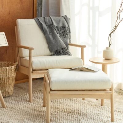 A wooden armchair with white cushions and a gray throw blanket is paired with a matching ottoman, an open book, a small round side table, and a vase on a textured rug.
