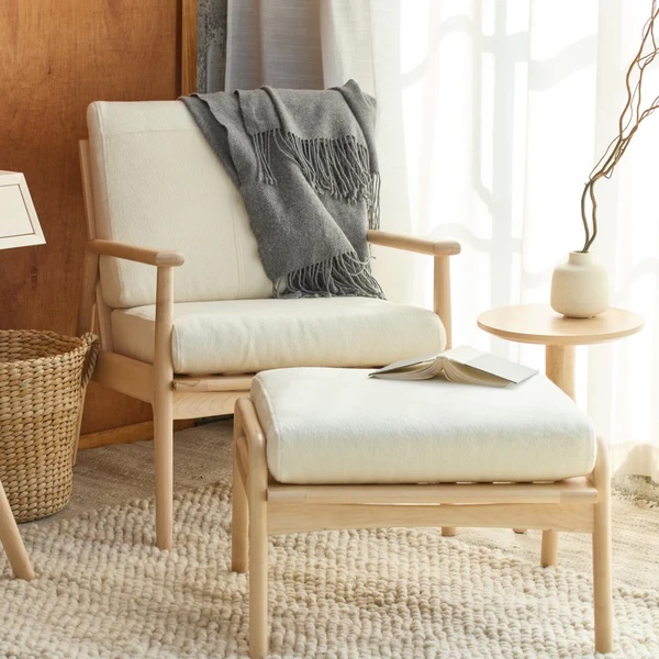 A wooden armchair with white cushions and a gray throw blanket is paired with a matching ottoman, an open book, a small round side table, and a vase on a textured rug.