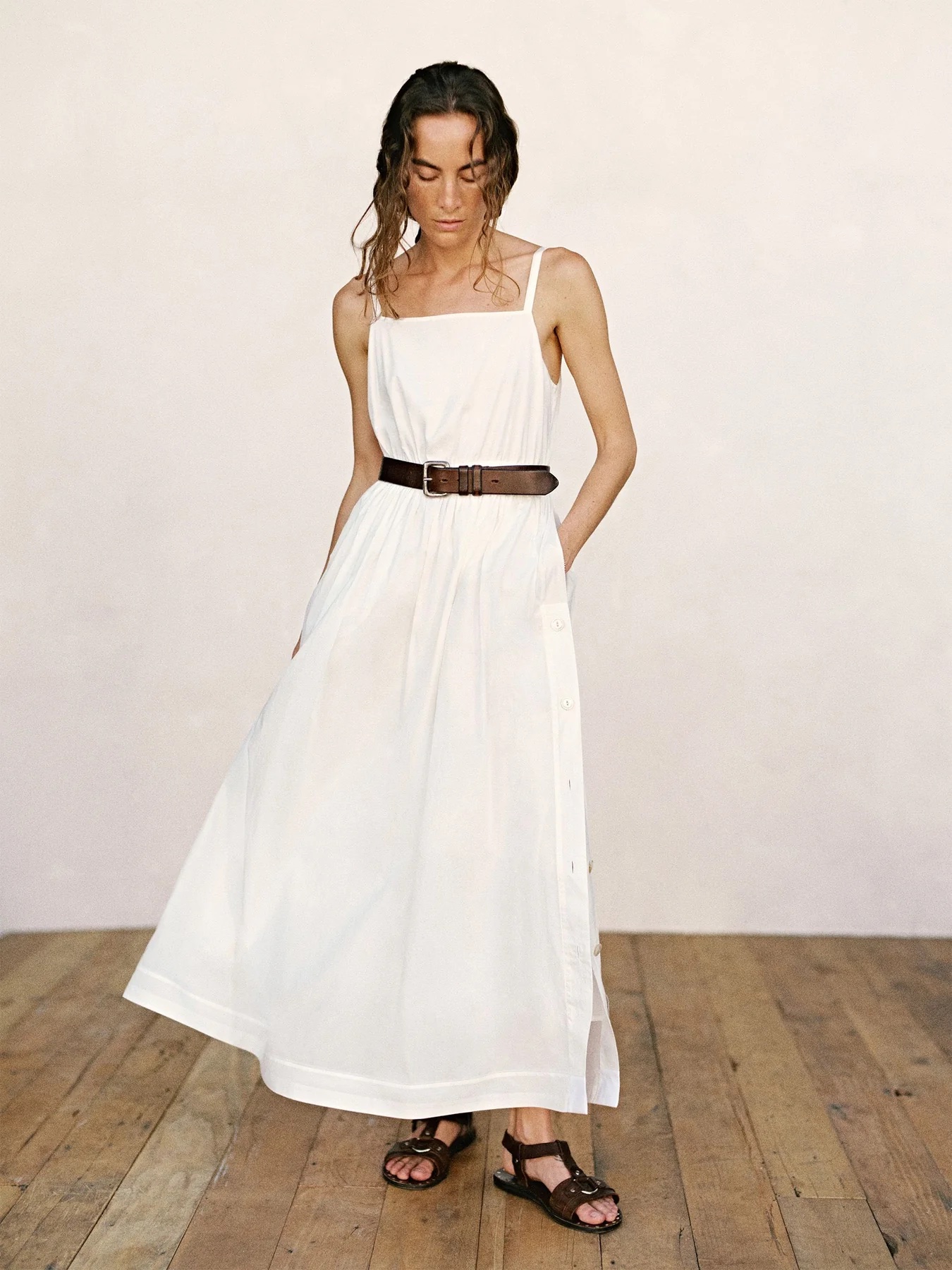 A woman wearing a long white sleeveless dress with a brown belt and brown sandals stands on a wooden floor against a plain light-colored wall.