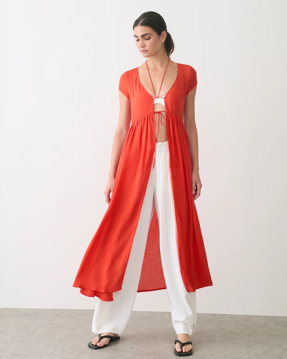A woman stands indoors wearing a red, short-sleeve, open-front dress over a white bralette and white wide-leg pants, paired with black sandals.