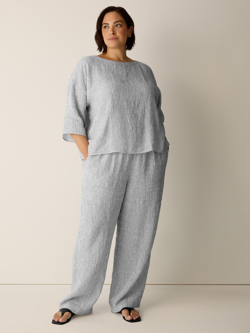 A woman stands against a plain background, wearing a loose-fitting, light gray textured top and matching pants with black sandals.