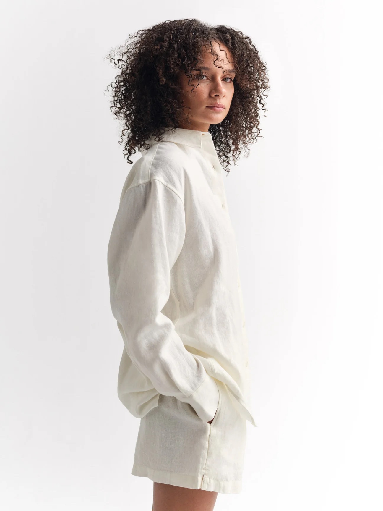 A person with curly hair wears a loose, long-sleeved, off-white button-up shirt and matching shorts, standing against a plain white background.