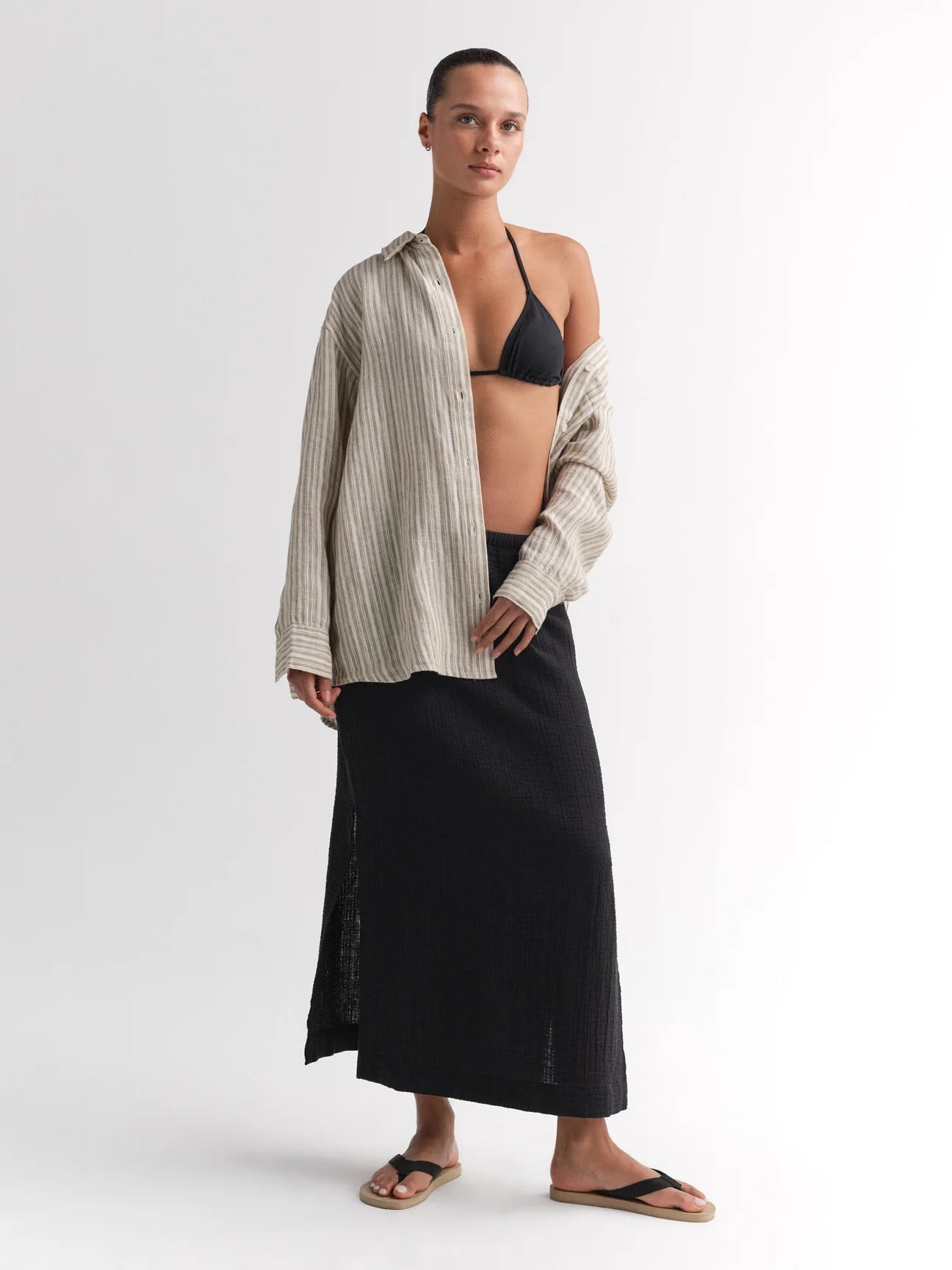 A person stands against a plain background wearing a black bikini top, an open beige striped shirt, a long black skirt, and black sandals.