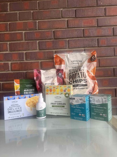 A selection of Thrive Market food products and teas, including chips, dried pineapple, pitted dates, fruit circles, teas, deluxe shells & cheese, and a small bottle, against a brick wall.
