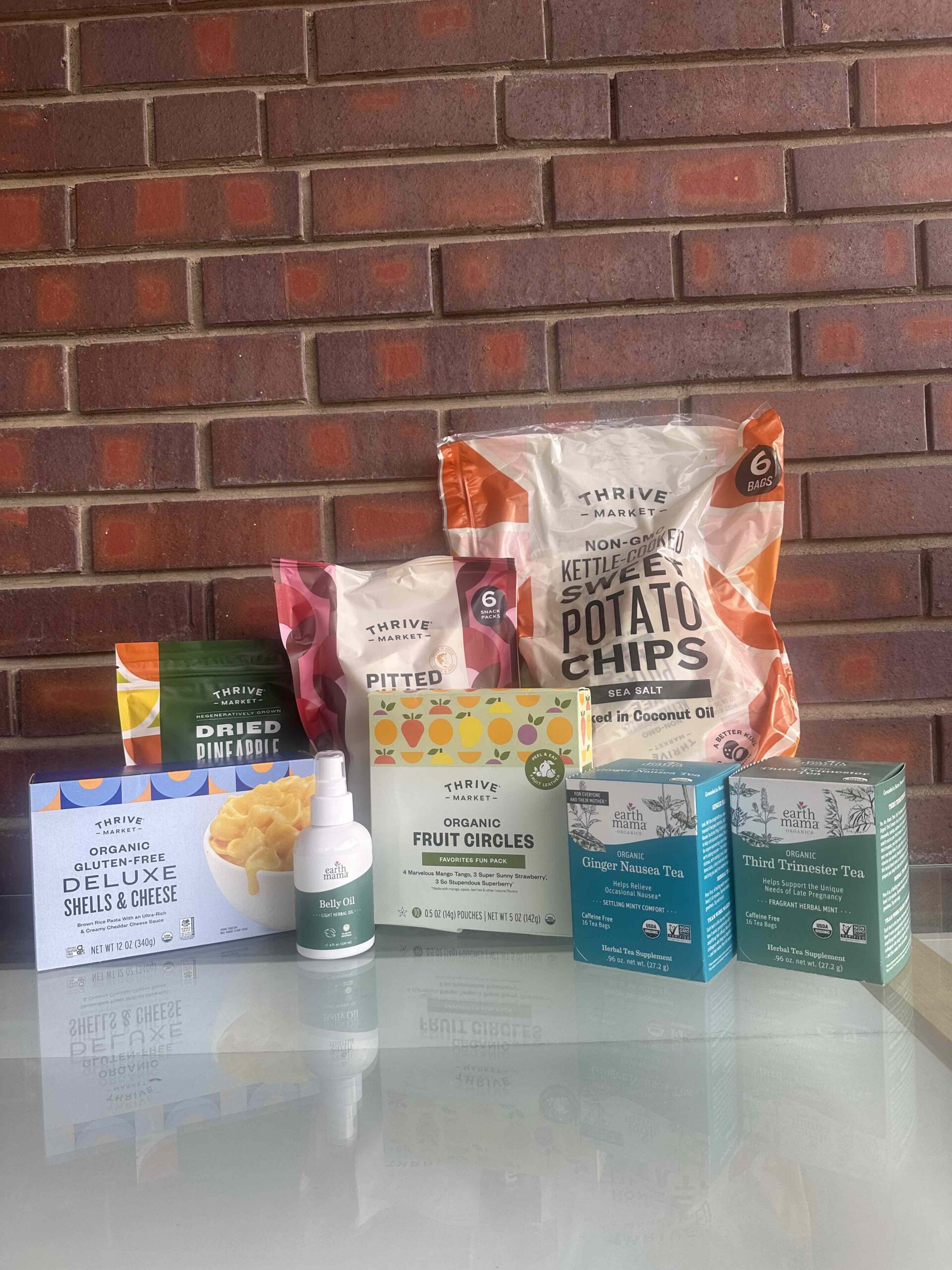 A selection of Thrive Market food products and teas, including chips, dried pineapple, pitted dates, fruit circles, teas, deluxe shells & cheese, and a small bottle, against a brick wall.