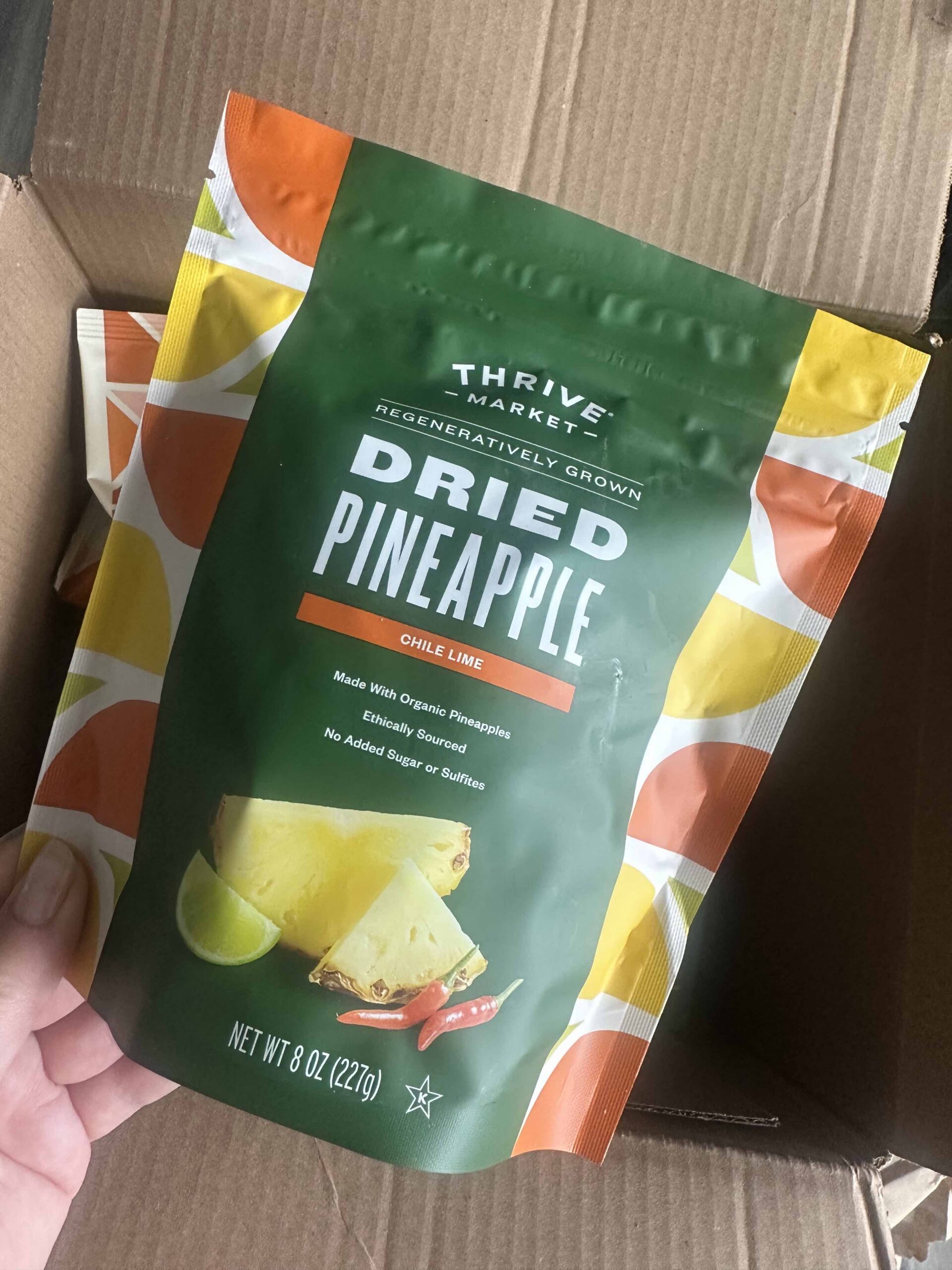 A hand holds a bag of Thrive Market regeneratively grown dried pineapple chili lime flavor, labeled as organic, ethically sourced, with no added sugar or sulfites.