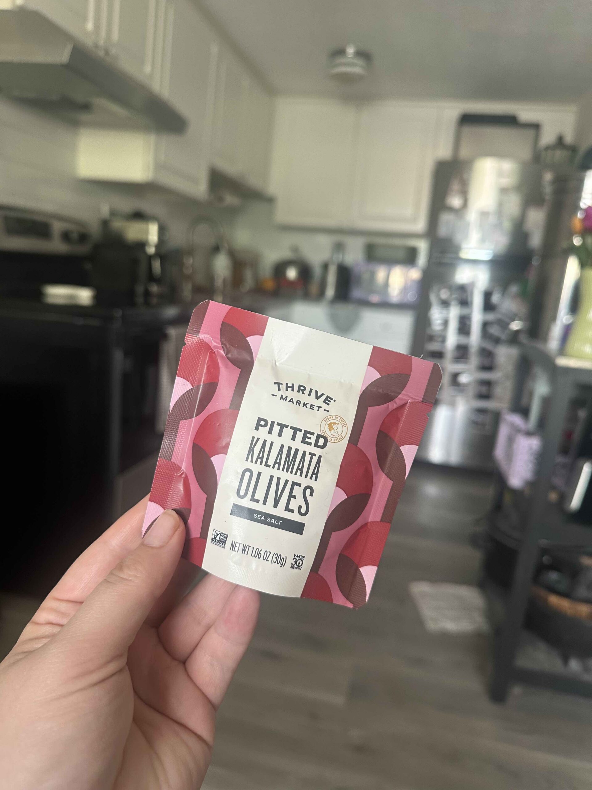 A hand holds a package of Thrive Market Pitted Kalamata Olives in a kitchen with white cabinets and various appliances in the background.