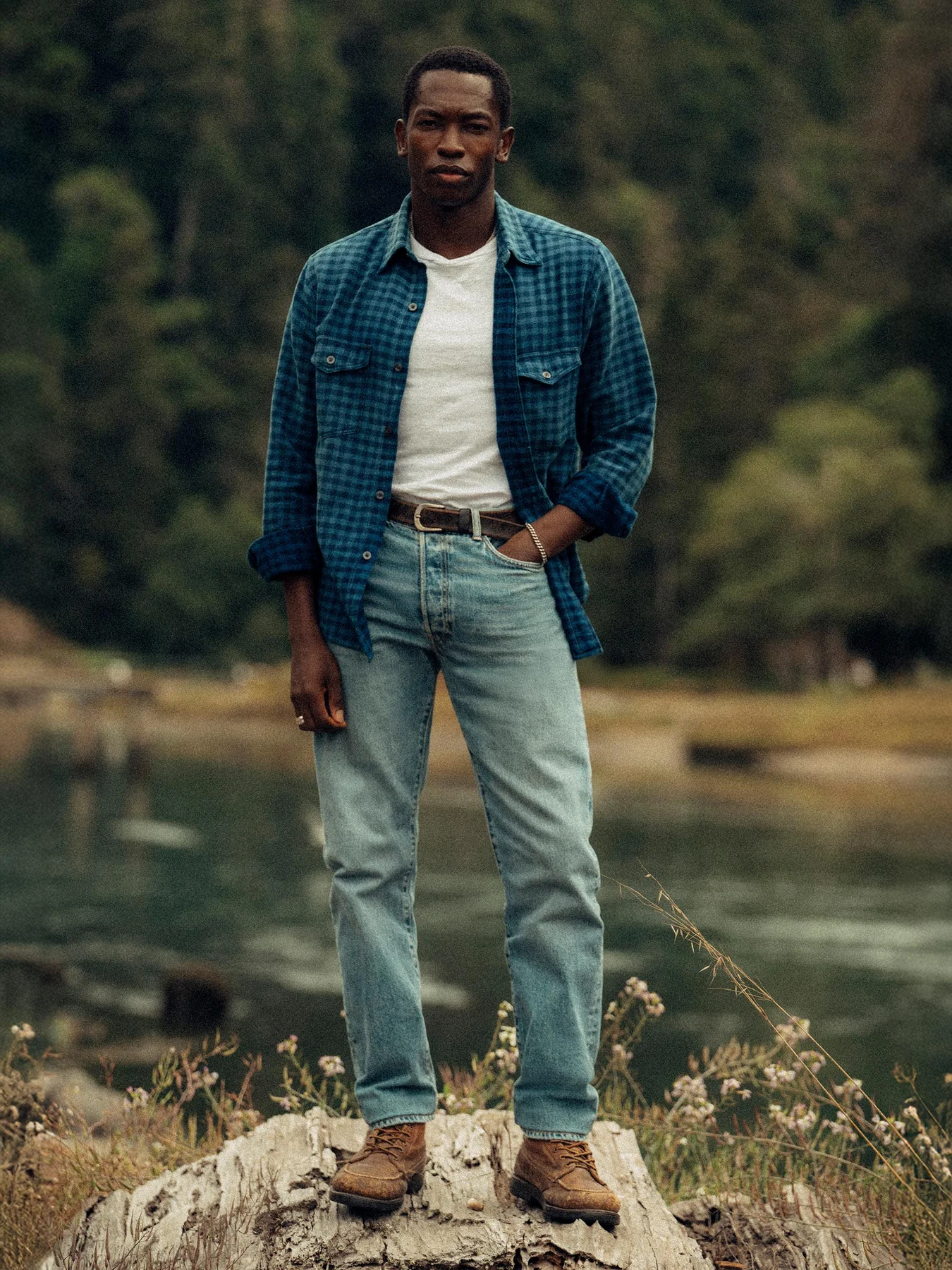 A man stands outdoors on a tree stump by a body of water, wearing a blue plaid shirt over a white t-shirt, light jeans, and brown boots, with a forested background.