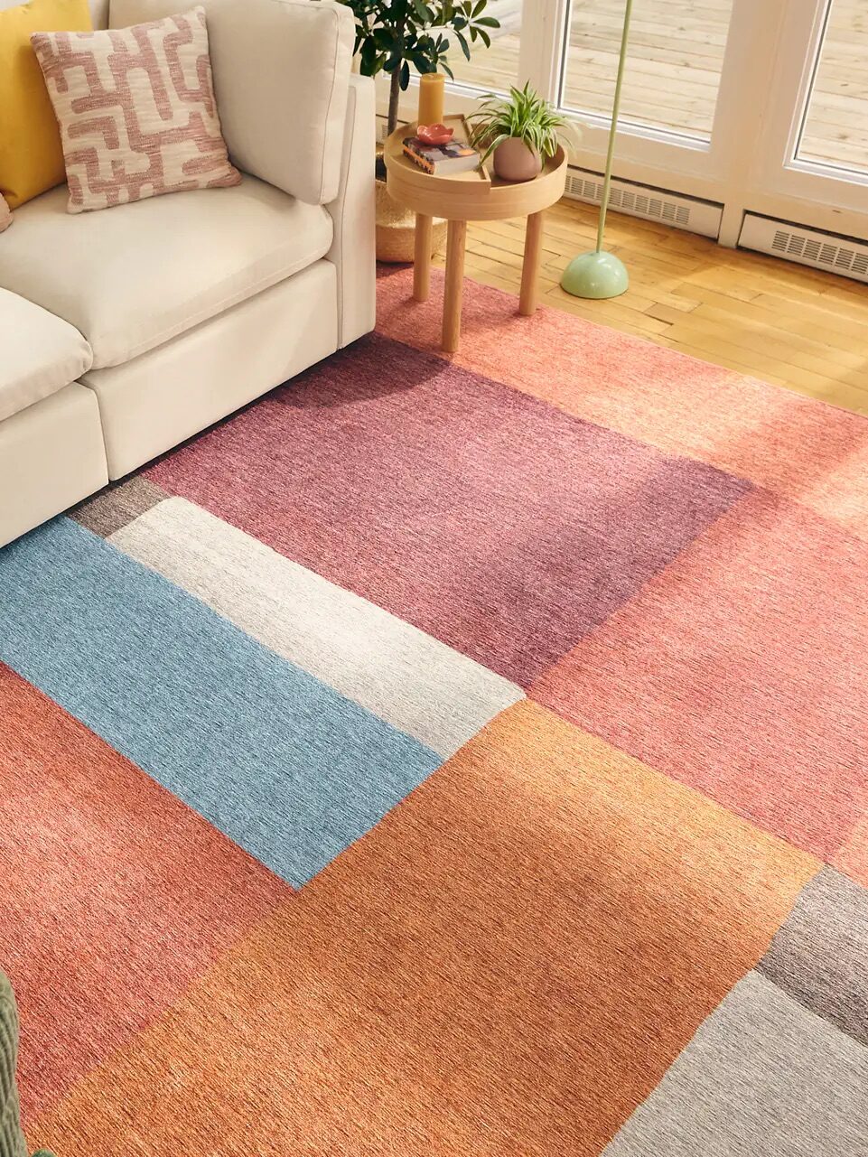 A living room with a white sofa, decorative pillows, a green chair, a plant, and a colorful geometric area rug on a wooden floor near large windows.