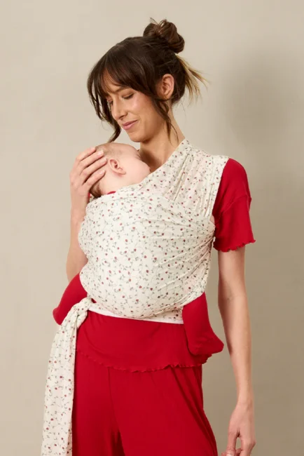 Woman in a red outfit holds a sleeping baby wrapped in a patterned white baby carrier against her chest.