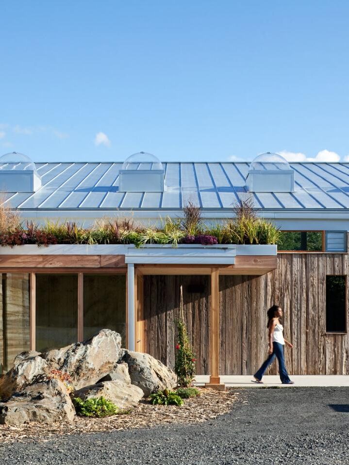 A person walks past a modern building with weathered wood siding, a metal roof with skylights, a planted roof overhang, and large decorative rocks by the entrance.