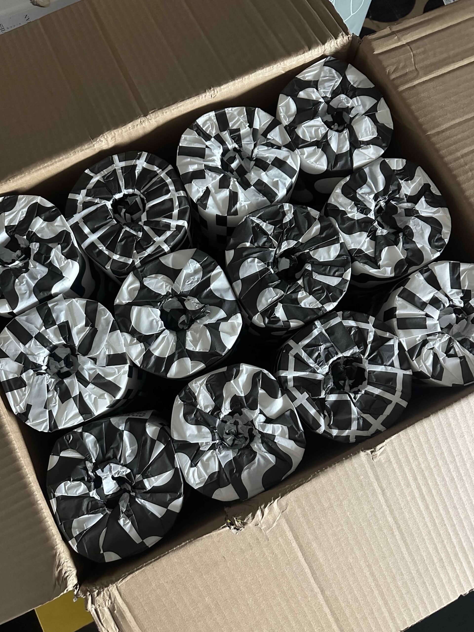 A cardboard box filled with twelve cylindrical items from Who Gives A Crap, each wrapped in black and white patterned paper.