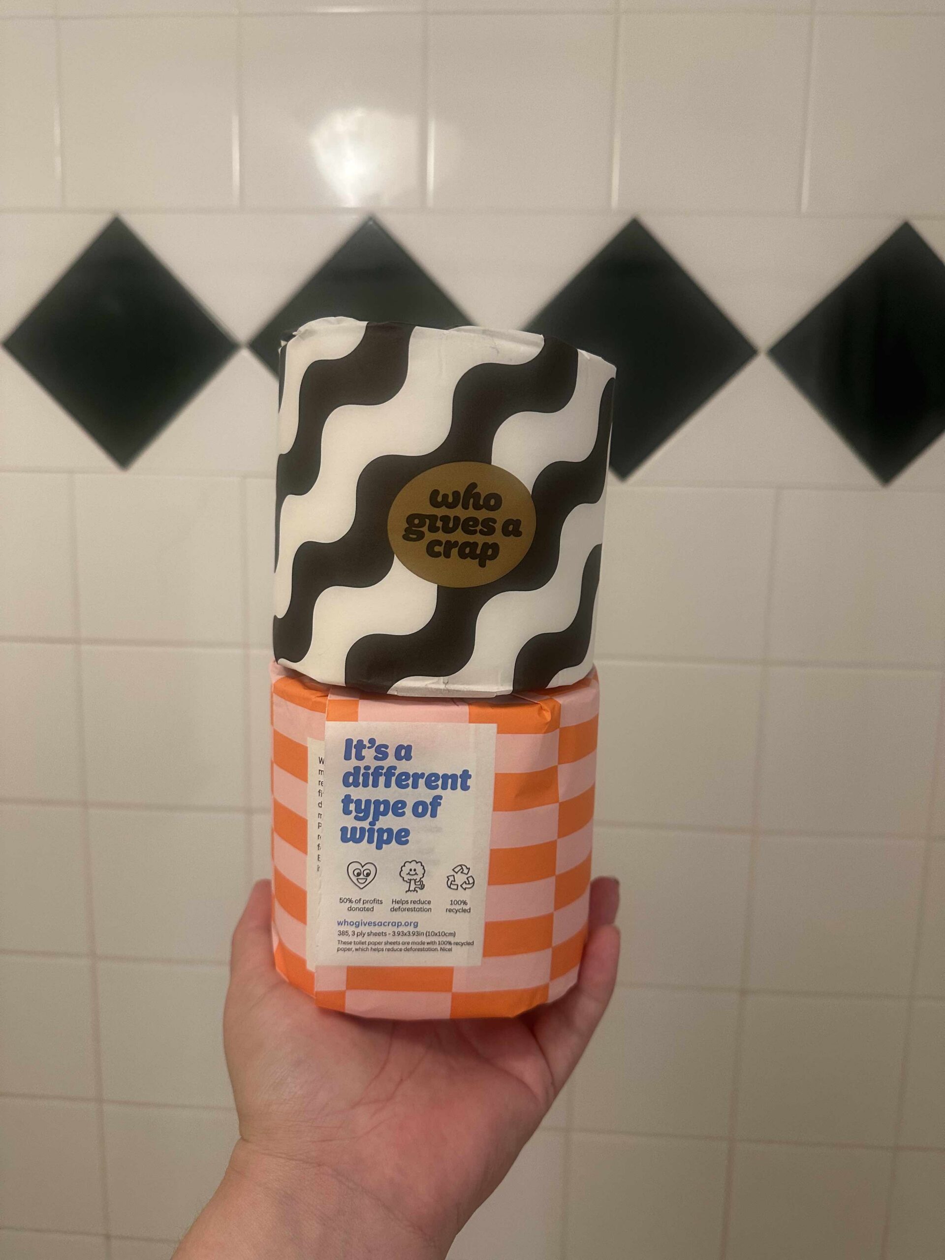 A hand holds a patterned toilet paper roll labeled "who gives a crap" above orange packaging that reads "It’s a different type of wipe," highlighting the eco-friendly ethos of Who Gives A Crap, against a tiled wall with dark green diamond accents.