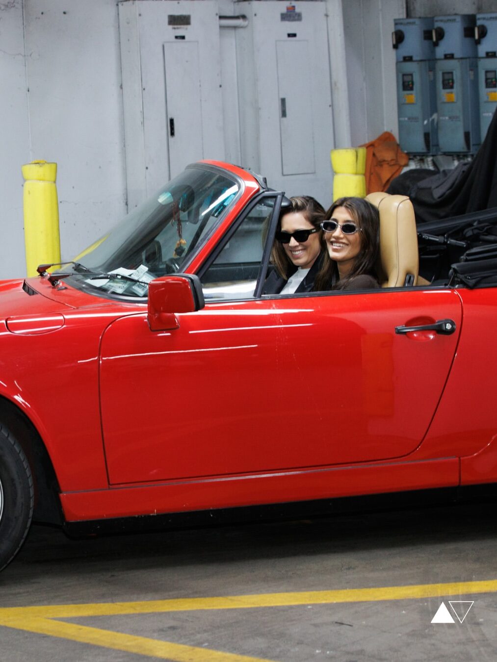 Two people wearing sunglasses sit in a red convertible car parked indoors, both looking toward the camera and smiling.