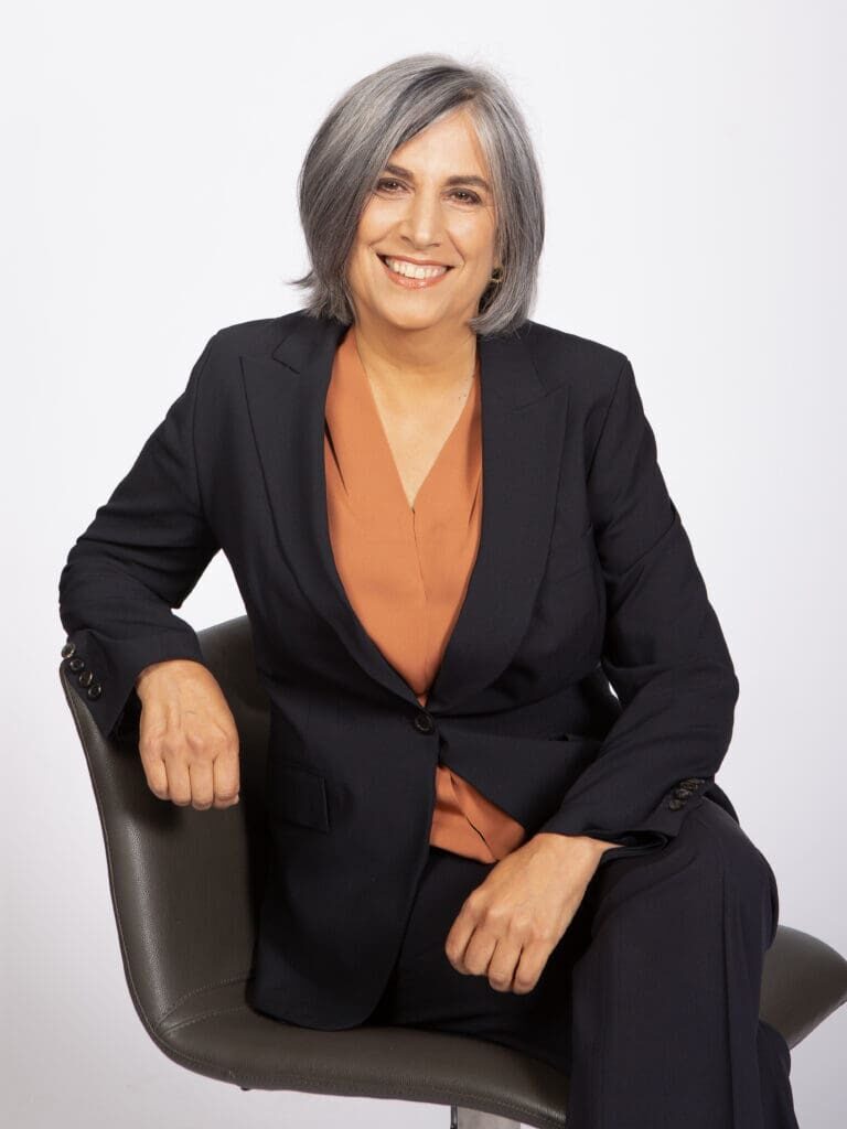 Woman with gray hair in a black suit and orange blouse sits on a chair, smiling at the camera against a plain white background.