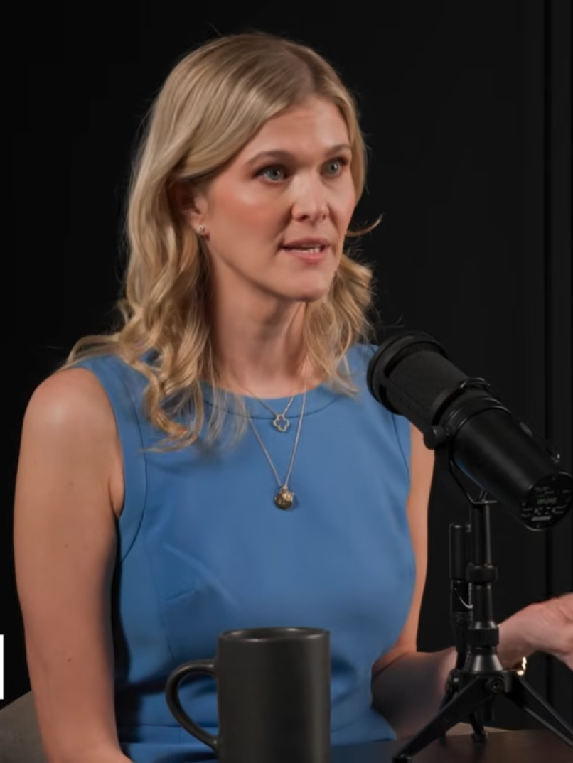 A woman with blonde hair wearing a blue sleeveless top speaks into a microphone during a podcast or interview. A mug and another microphone are visible on the table.