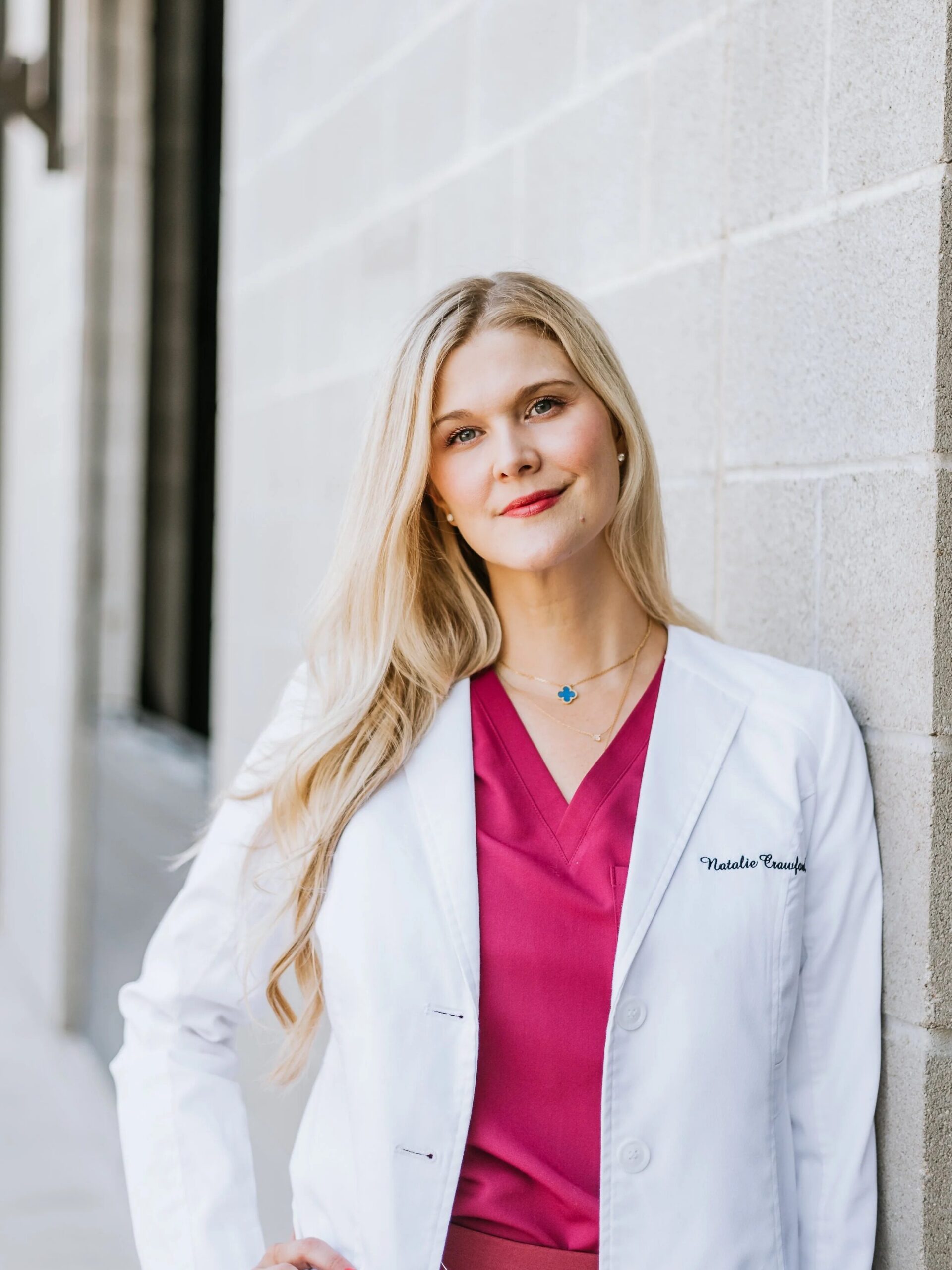A woman in a white lab coat over magenta scrubs stands against a light-colored brick wall, looking at the camera.