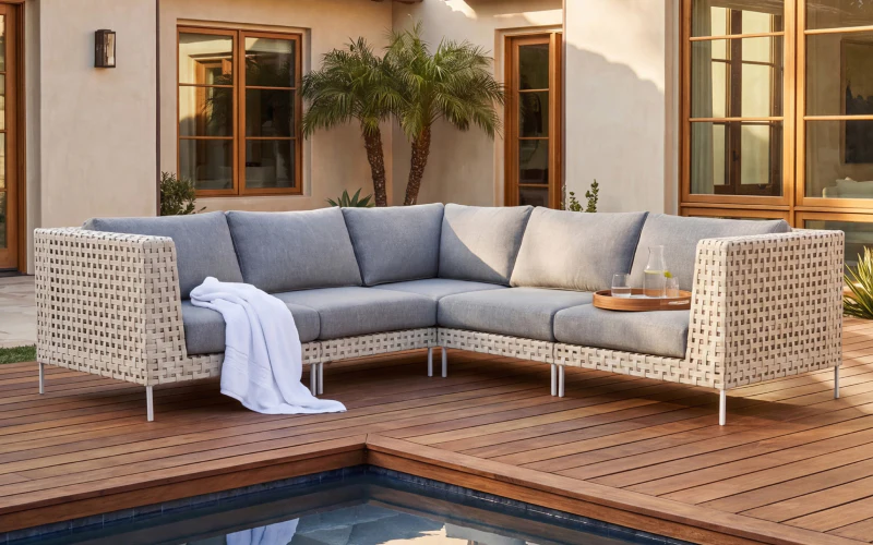 A light gray outdoor sectional sofa with a white blanket and a wooden tray holding drinks sits on a wooden deck near a pool, with a modern house and palm tree in the background.