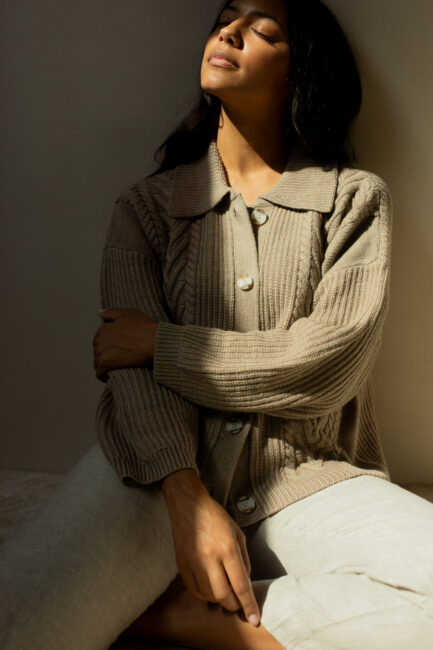 A person sits with eyes closed, head tilted back, and sunlight illuminating their face, wearing a beige knit sweater and light-colored pants against a plain background.