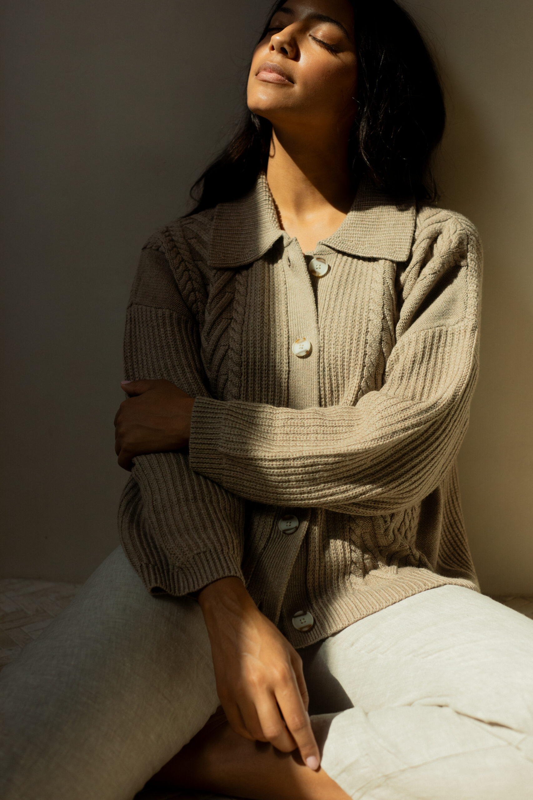 A person sits with eyes closed, head tilted back, and sunlight illuminating their face, wearing a beige knit sweater and light-colored pants against a plain background.