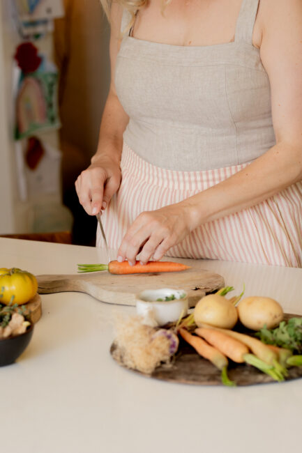 A woman in a beige apron slices a carrot on a wooden cutting board with potatoes and other vegetables nearby on a kitchen counter.