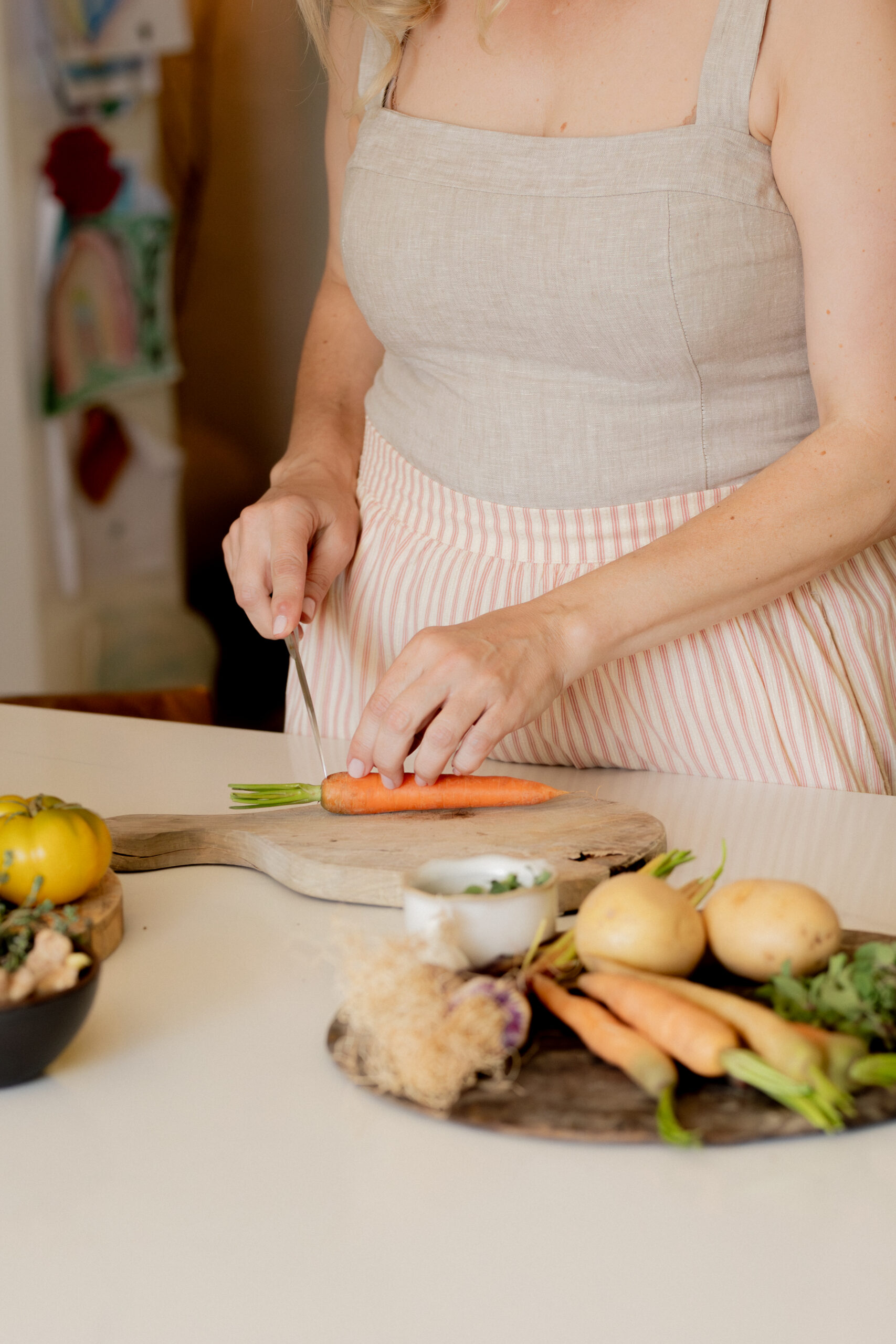A woman in a beige apron slices a carrot on a wooden cutting board with potatoes and other vegetables nearby on a kitchen counter.