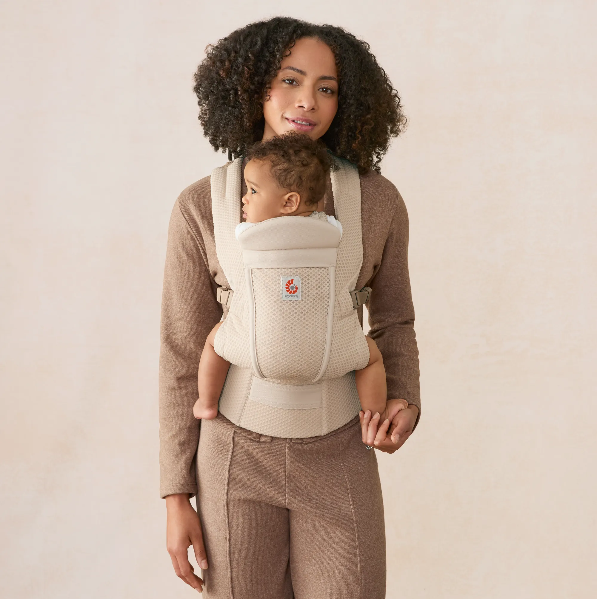 Woman wearing a brown outfit holds a baby in a beige front-facing baby carrier, standing against a plain light background.