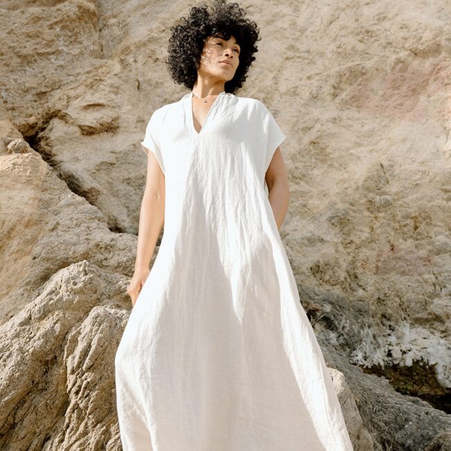 A person with curly hair stands on rocky terrain wearing a loose, white, short-sleeved dress.