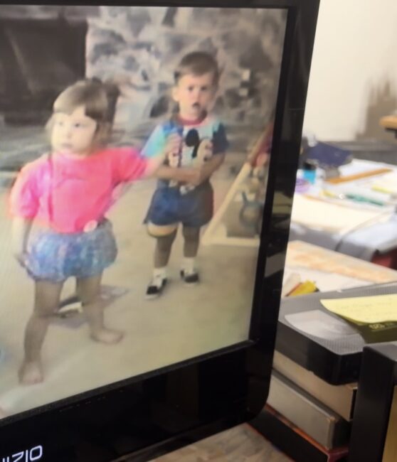 A young girl and boy stand indoors, the girl in a pink shirt and denim skirt, the boy in a Mickey Mouse shirt and shorts. The image is displayed on a television screen.