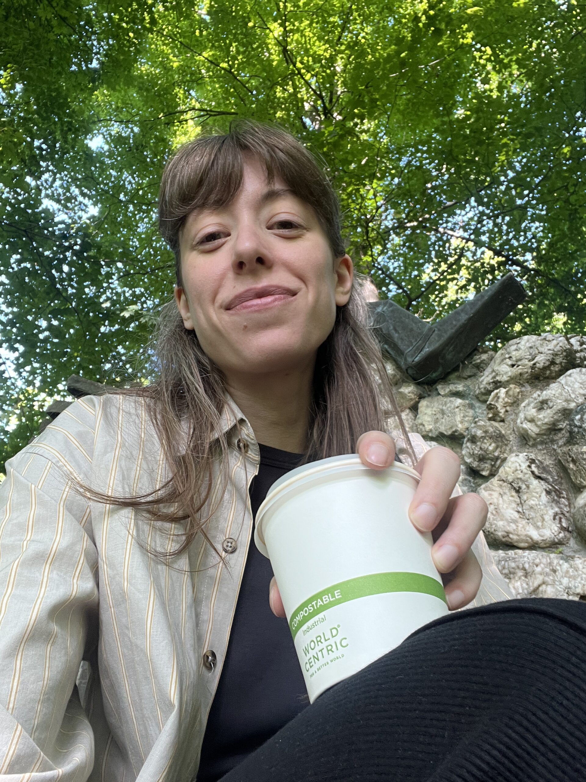 A person with long brown hair holds a compostable cup and smiles at the camera while sitting outside near rocks and green trees.