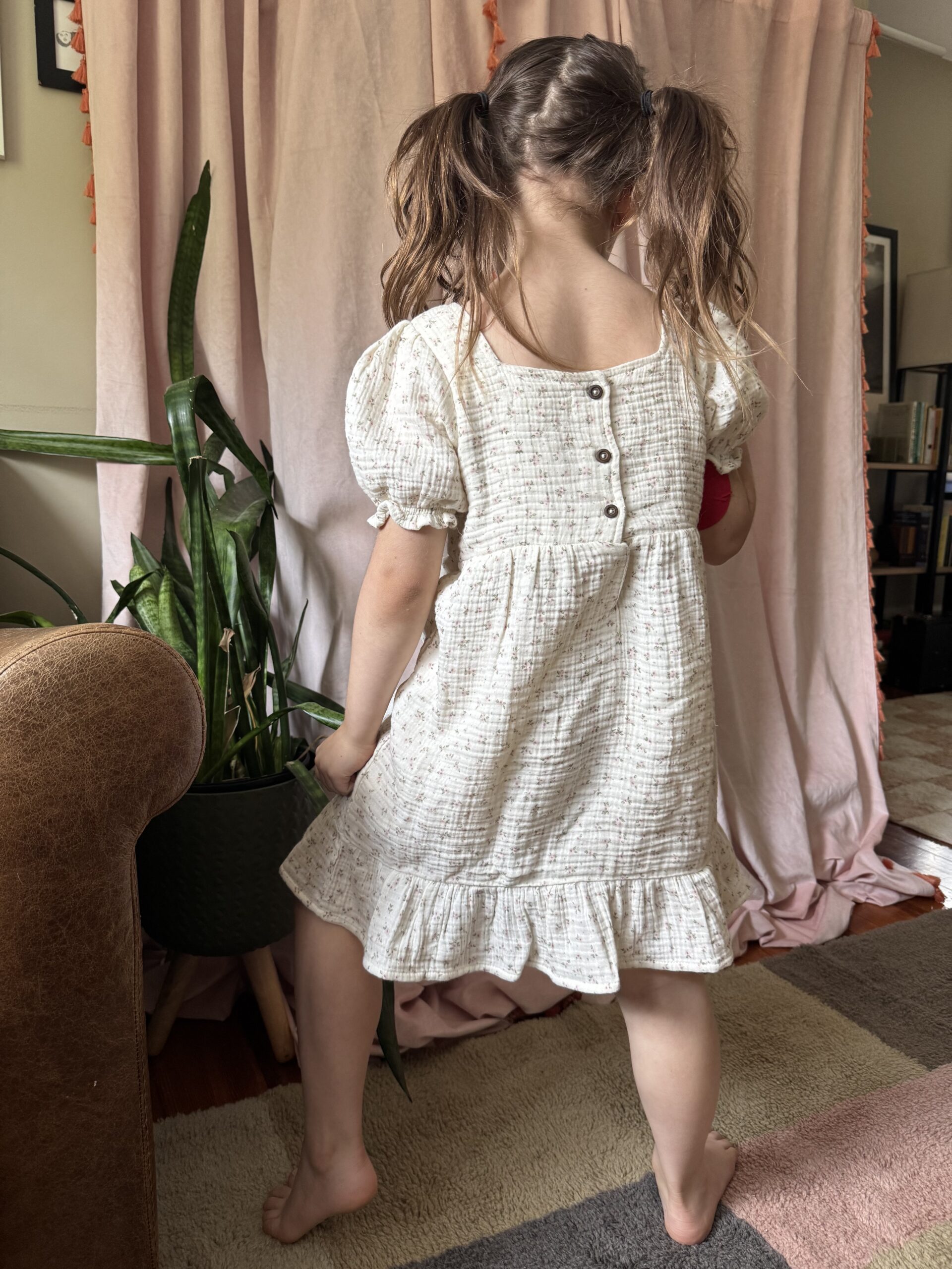 A young girl with pigtails stands barefoot on a rug, facing away, wearing a light-colored dress with buttons on the back. There is a potted plant and curtains in the background.