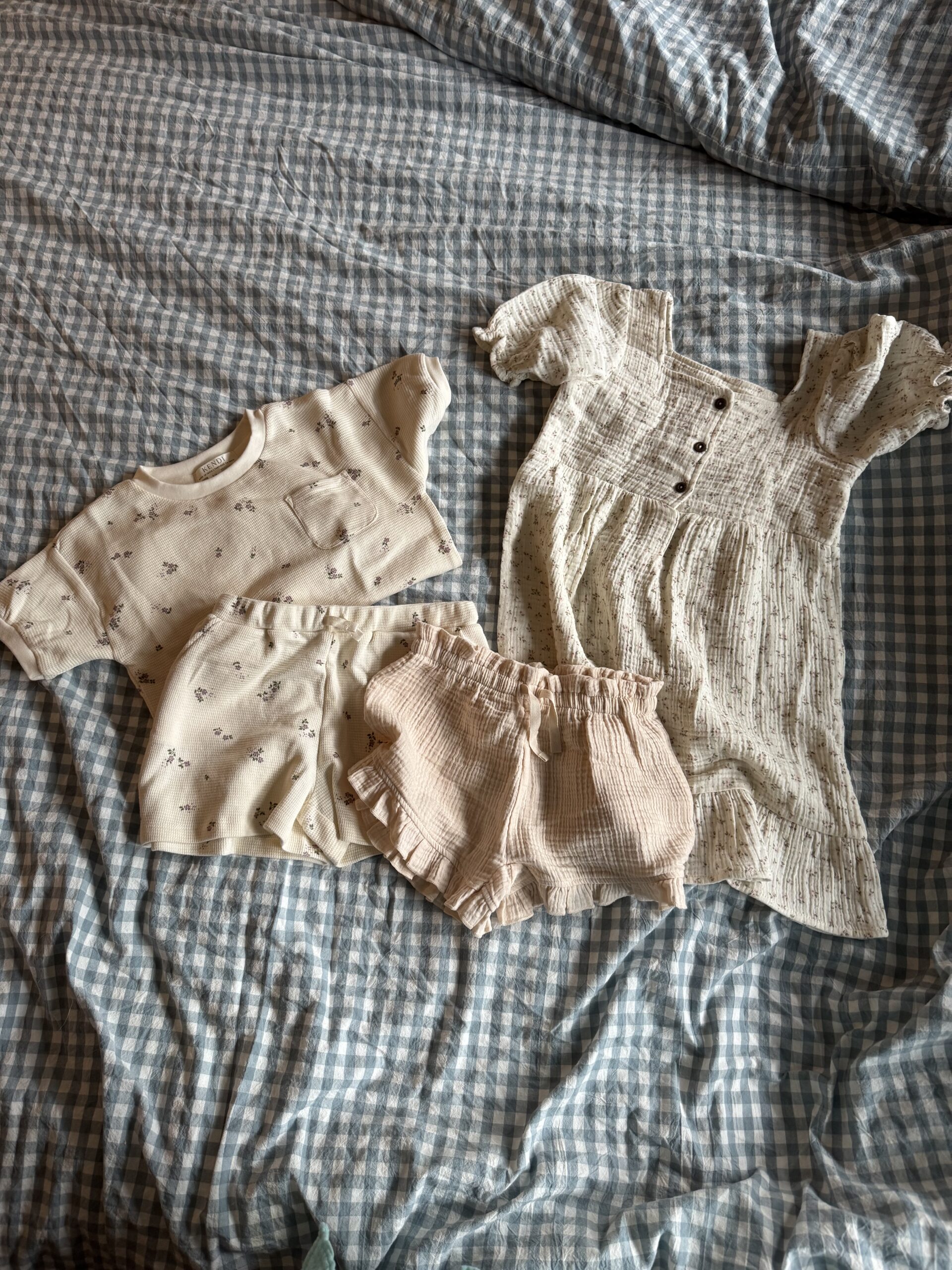 Three pieces of children's clothing—a short-sleeve shirt, a pair of shorts, and a short-sleeve dress—are laid out on a blue and white checkered bedspread.