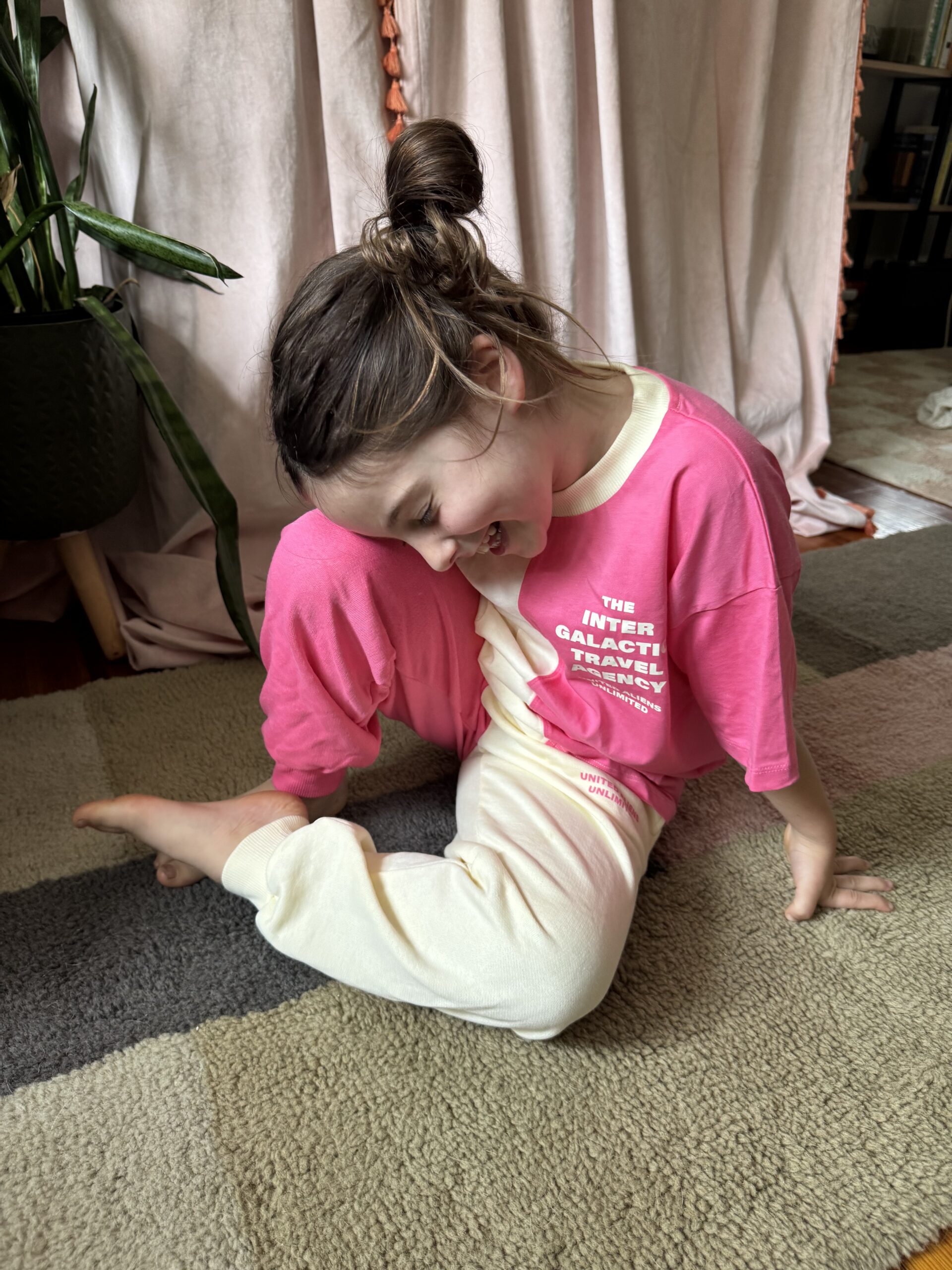 A child in pink and cream pajamas sits on a carpeted floor, smiling and looking down with one leg crossed and a plant in the background.