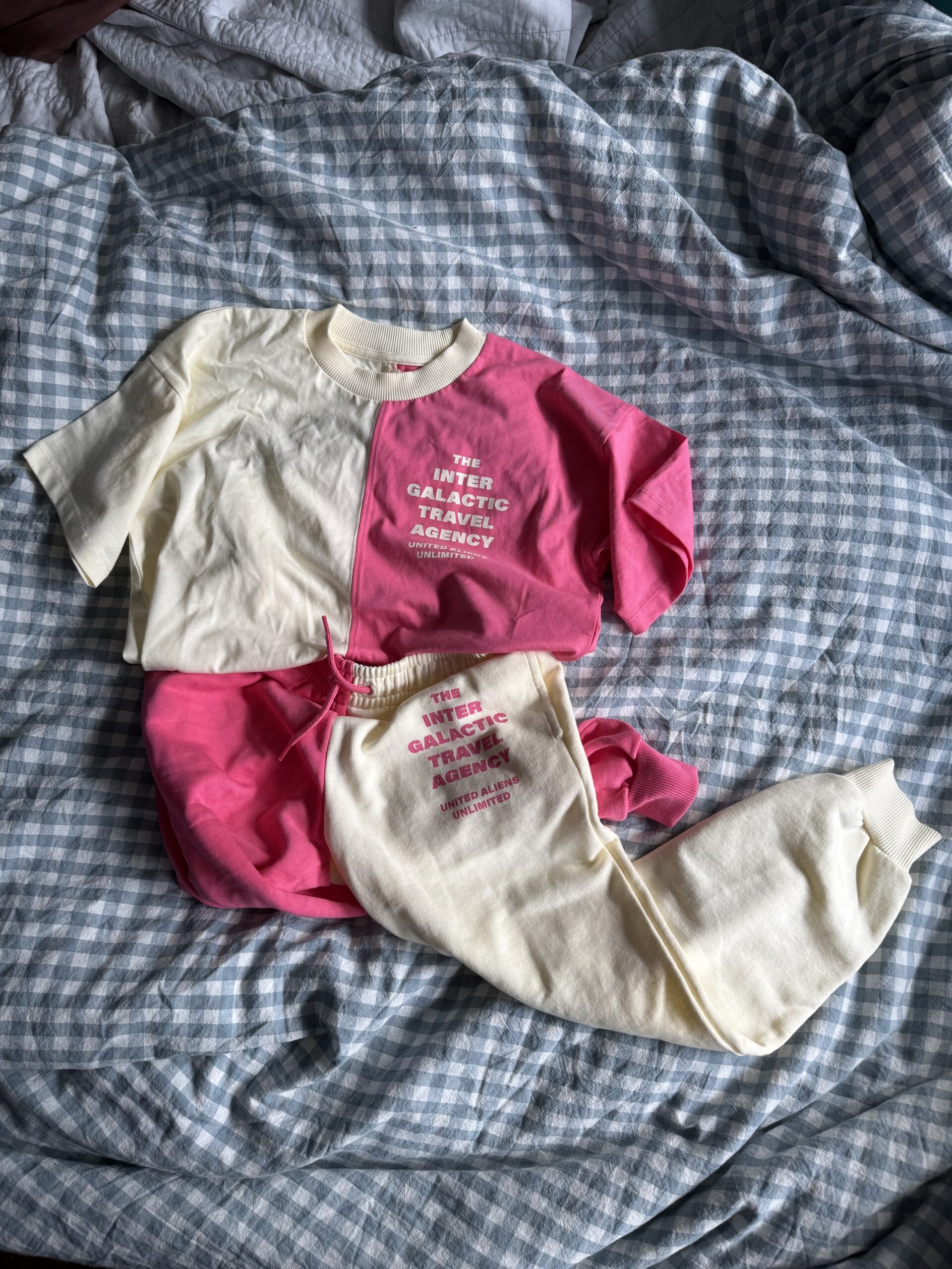 Two-toned pink and cream t-shirt and sweatpants with "The Inter Galactic Trading Agency" text, laid out on a blue and white plaid bedspread.