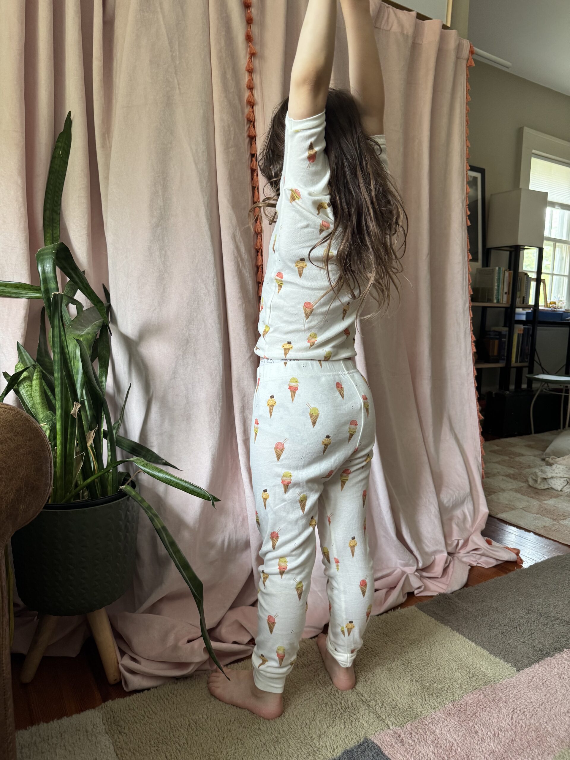 A child in ice cream cone pajamas stretches with arms raised, standing next to a potted plant in a living room with pink curtains.