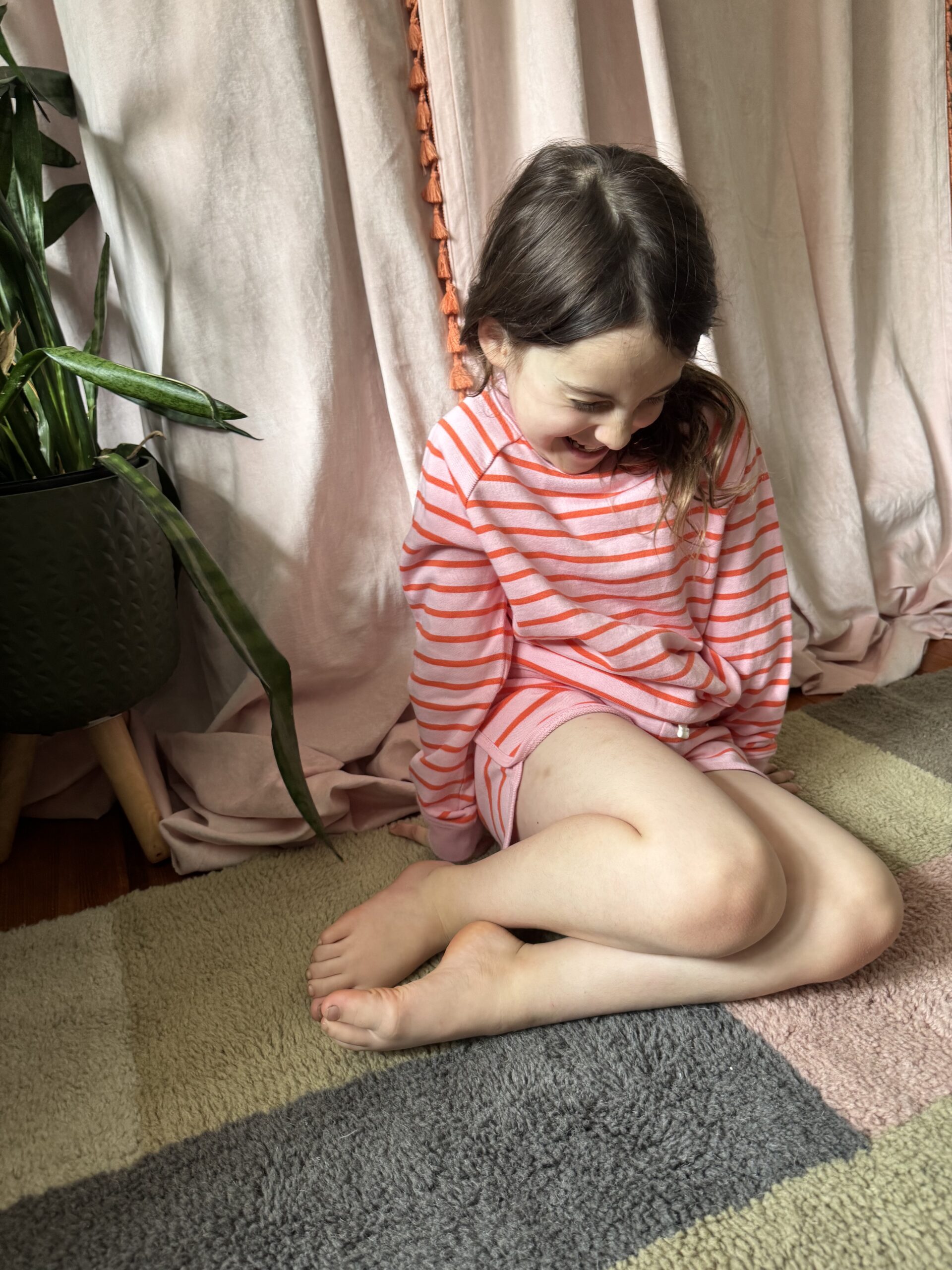 A young girl in a pink and orange striped outfit sits cross-legged on a rug, looking down and smiling, with a plant and light-colored curtains in the background.