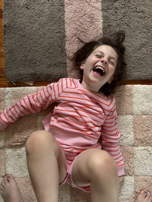 A child in a pink and red striped shirt laughs while lying on a checkered rug, with an adult's feet partially visible at the bottom of the image.
