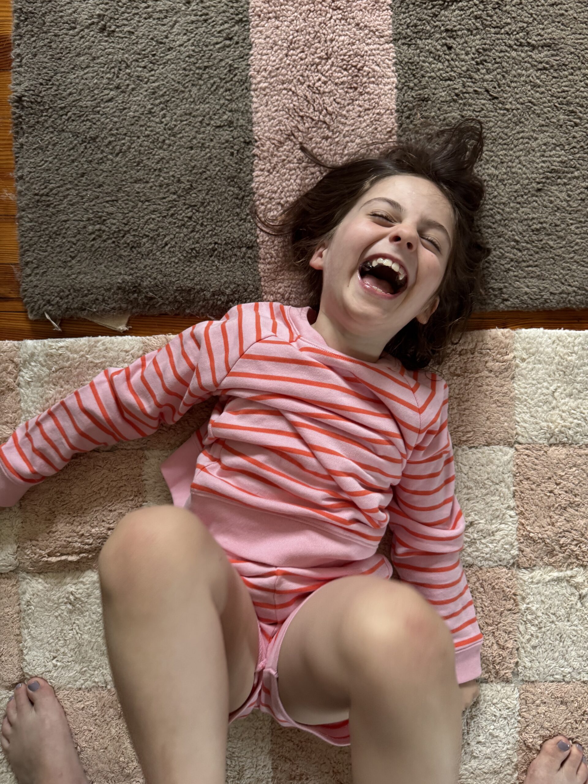 A child in a pink and red striped shirt laughs while lying on a checkered rug, with an adult's feet partially visible at the bottom of the image.