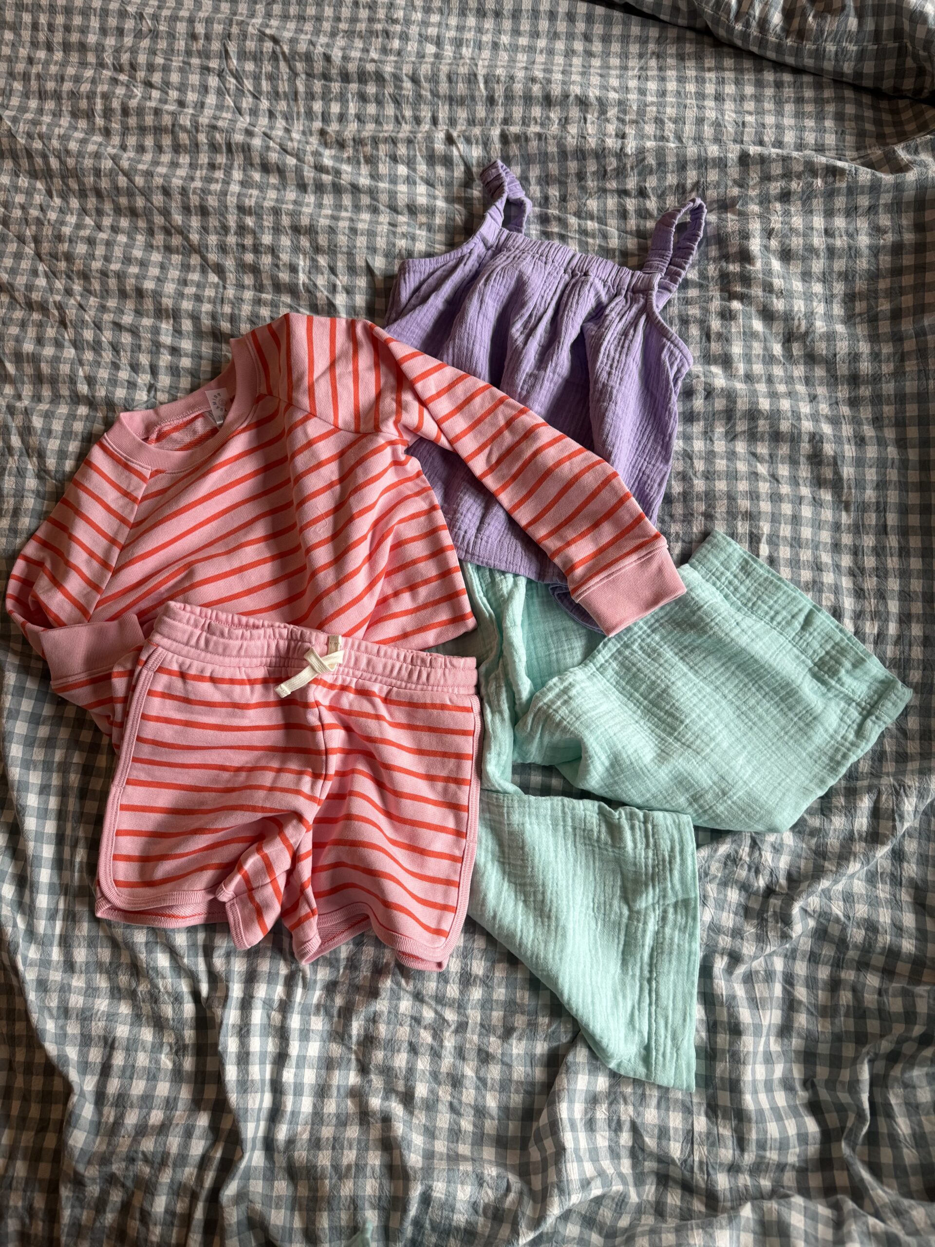 A set of children's clothes on a checkered bedspread: a pink and red striped long-sleeve shirt, matching shorts, a lilac tank top, and light aqua pants.
