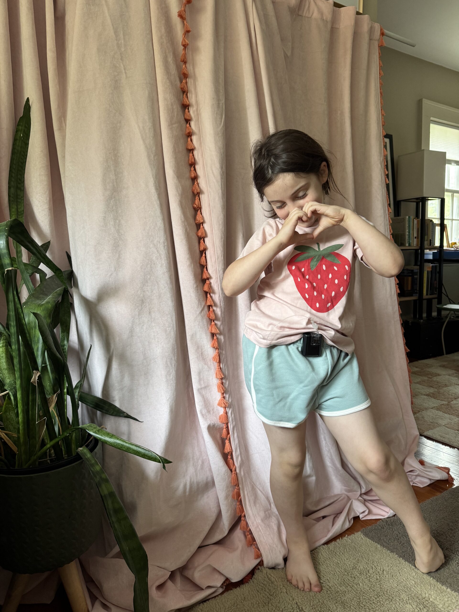 A child in a strawberry t-shirt and blue shorts stands indoors, making a heart shape with their hands in front of a pink curtain and a potted plant.