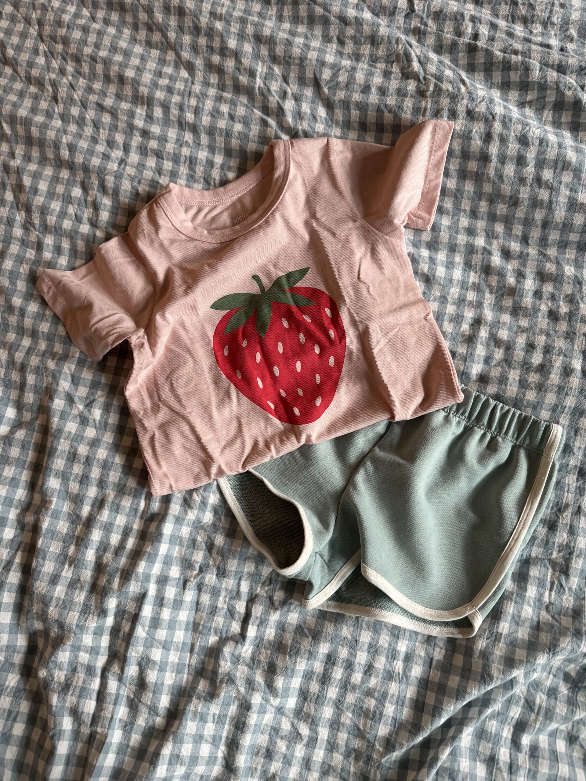 A child’s outfit with a pink T-shirt featuring a large strawberry graphic and a pair of green shorts, laid out on a blue and white checkered fabric.