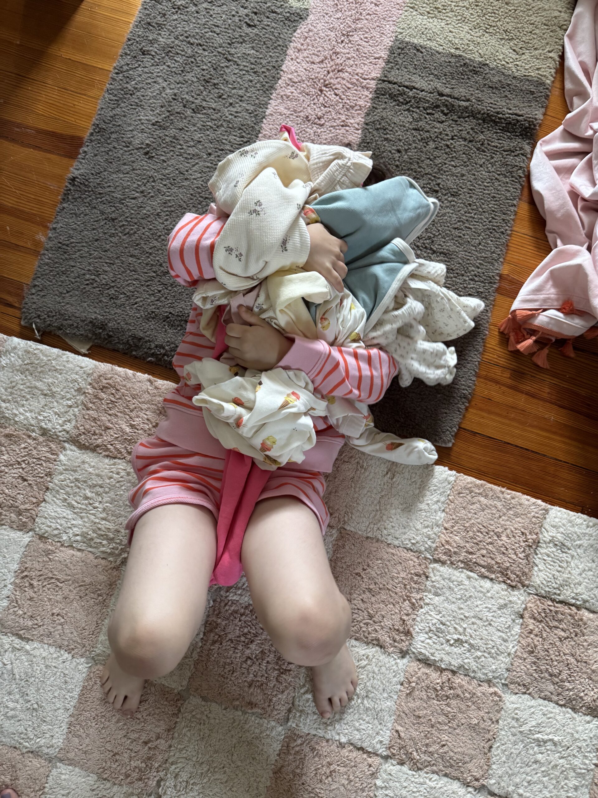 A child in pink pajamas lies on a rug, holding a large pile of clothes and blankets over their face.