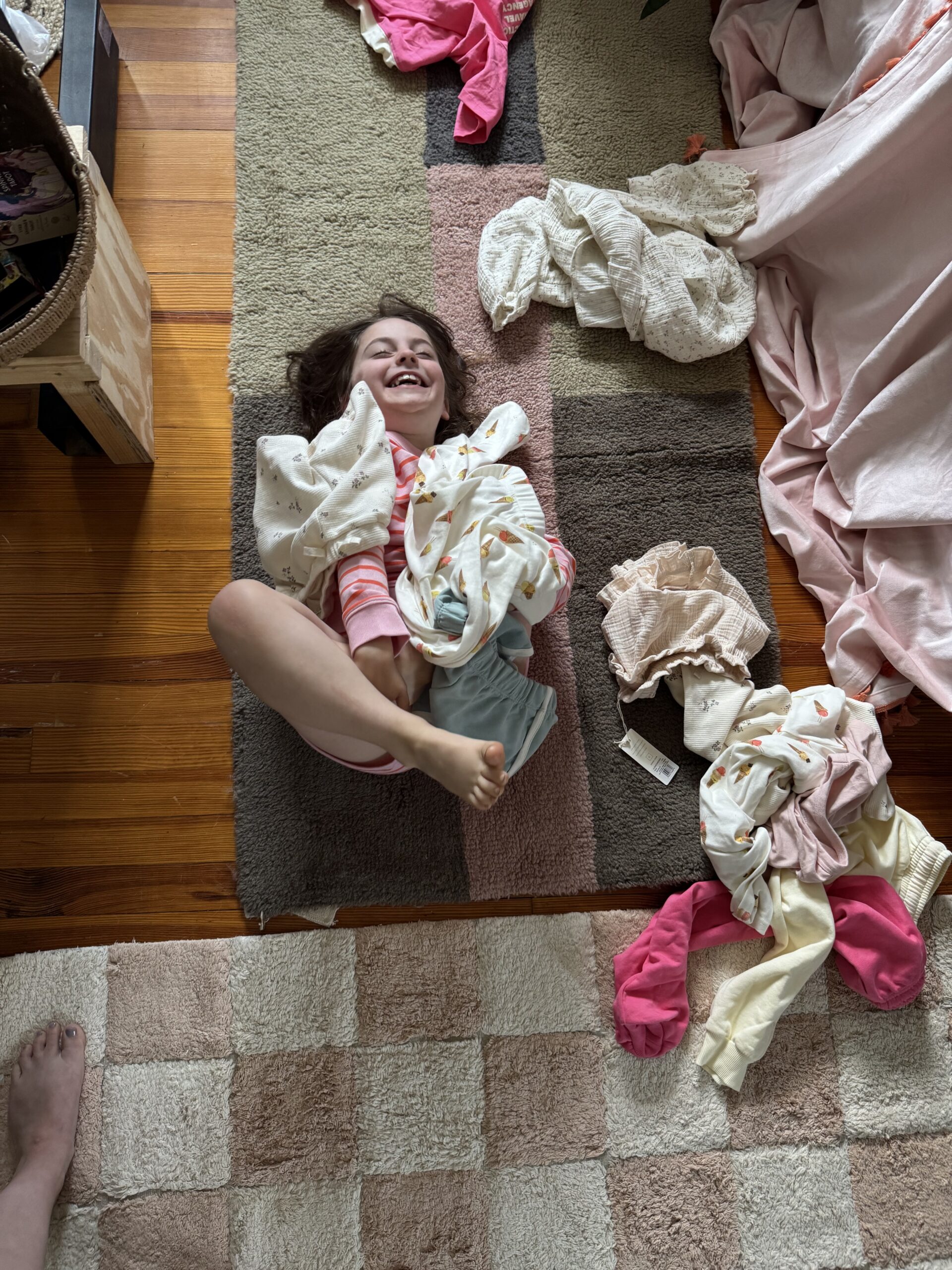 Young girl lying on a rug surrounded by scattered clothes and bedding, smiling and holding a blanket, with an adult’s foot visible in the lower left corner.