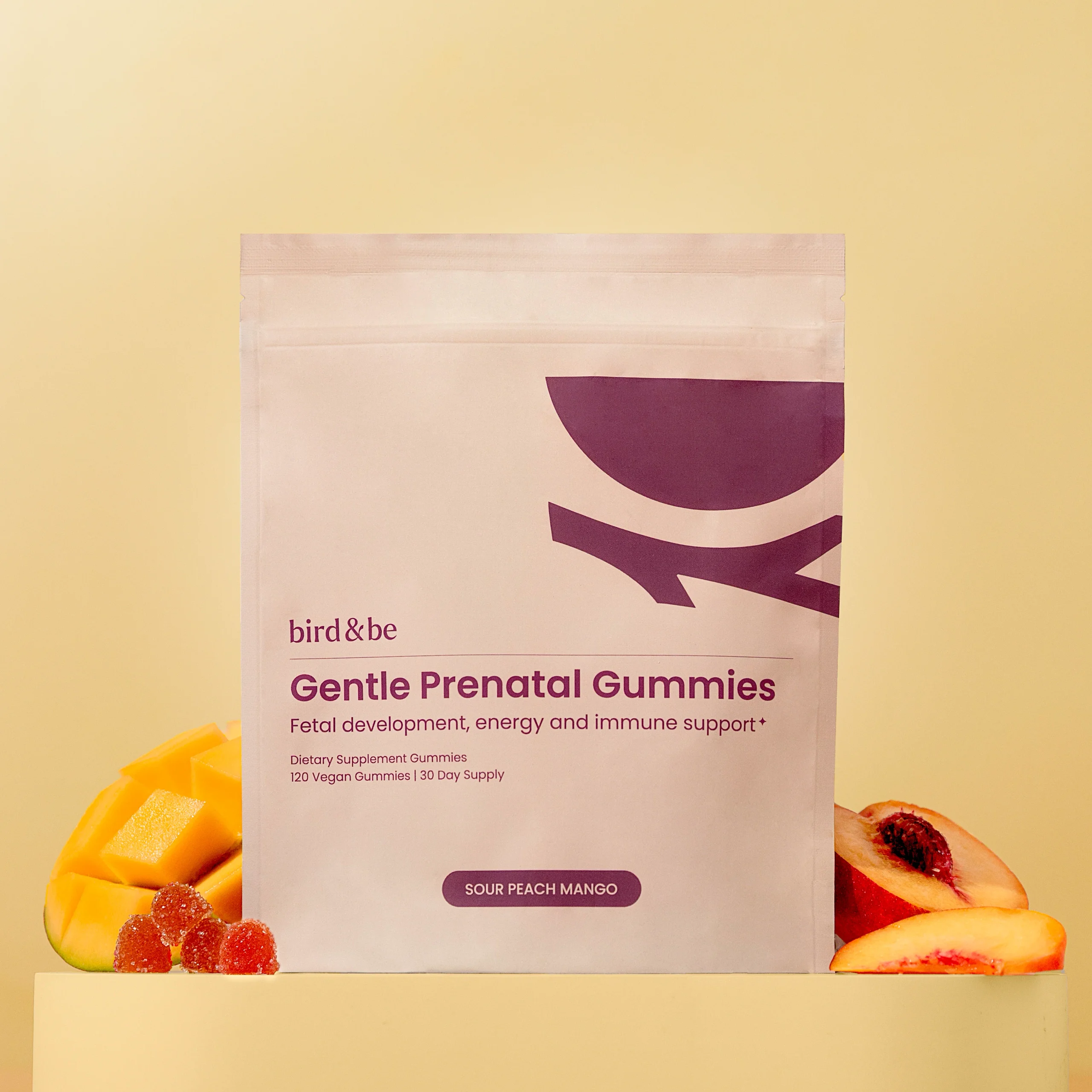 A package of bird&be Gentle Prenatal Gummies is displayed with sliced mango and peach pieces, against a beige background.