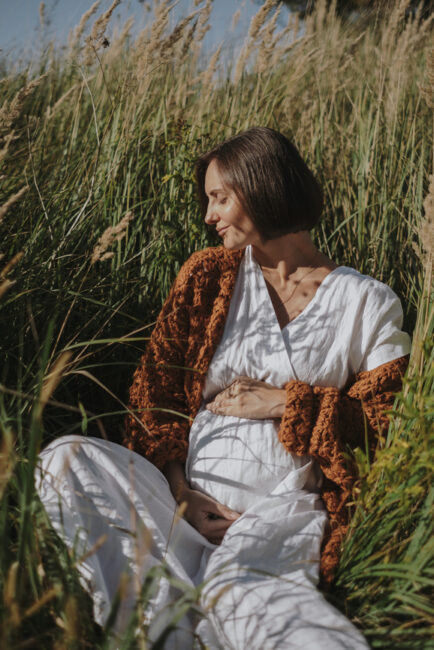 A woman in a white outfit and brown shawl sits in tall grass, gently holding her pregnant belly and looking down.