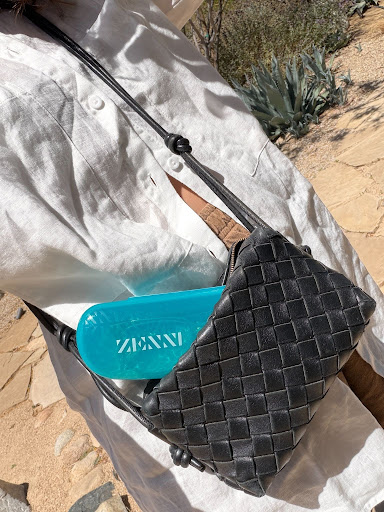 Person wearing a white button-up shirt carries a black woven crossbody bag with a bright blue rectangular object partially visible inside.